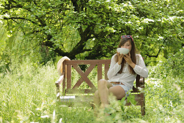 Junge Frau trinkt auf einer Holzbank in einem Garten - LAF000893