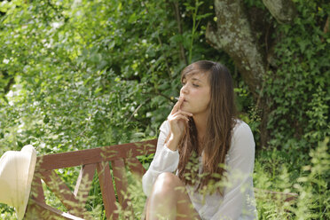 Young woman smoking on wooden bench in a garden - LAF000895