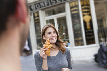 France, Paris, portrait of young woman showing croissant in front of pastry shop - FMKF001352