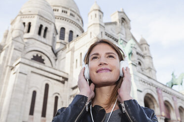Frankreich, Paris, Porträt einer glücklichen jungen Frau, die mit Kopfhörern vor der Sacre Coeur Musik hört - FMKF001261