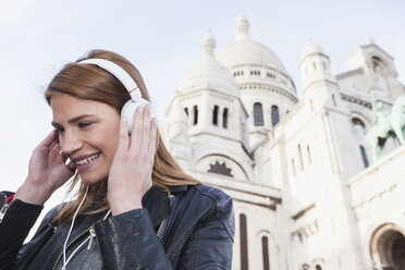 Frankreich, Paris, Porträt einer glücklichen jungen Frau, die mit Kopfhörern vor der Sacre Coeur Musik hört - FMKF001262