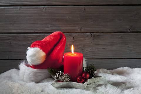 Angezündete rote Kerze und Weihnachtsmütze auf Kunstschnee vor grauer Holzwand, lizenzfreies Stockfoto