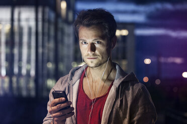 Porträt eines jungen Mannes mit Smartphone und Kopfhörern beim Musikhören in der Nacht - RBF001798