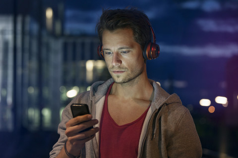 Porträt eines jungen Mannes mit Smartphone und Kopfhörern beim Musikhören in der Nacht, lizenzfreies Stockfoto