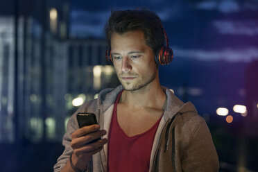 Porträt eines jungen Mannes mit Smartphone und Kopfhörern beim Musikhören in der Nacht - RBF001799