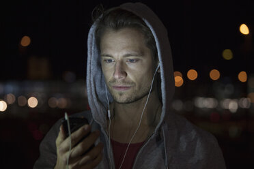 Portrait of young man wearing hooded jacket listening music at night - RBF001803