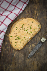 Slice of bread with obazda sprinkled with chives, knife and cloth on dark wood - LVF001433
