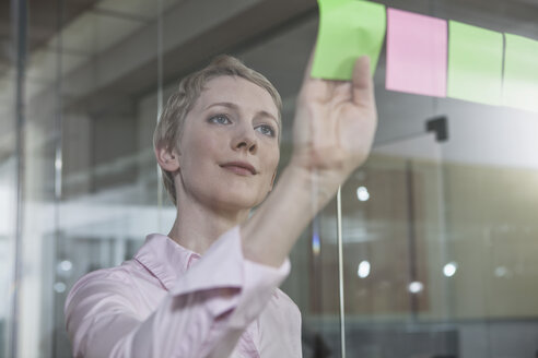 Germany, Munich, Businesswoman in office, putting sticky notes on glass pane - RBYF000517