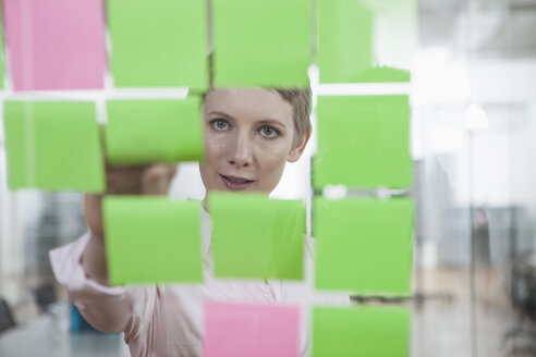Germany, Munich, Businesswoman in office, putting sticky notes on glass pane - RBYF000518