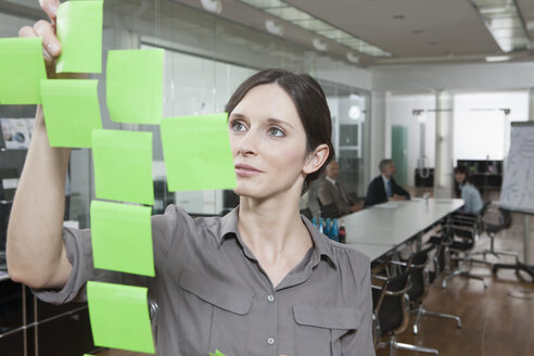 Germany, Munich, Businesswoman in office, putting sticky notes on glass pane - RBYF000622