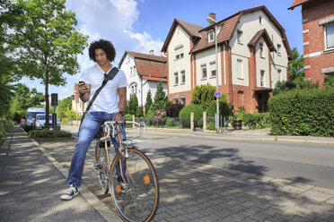 Junger männlicher Student sitzt auf einem Fahrrad und schaut auf sein Smartphone - VTF000284