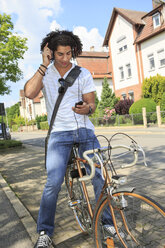 Junger männlicher Student mit Kopfhörern, der auf einem Fahrrad sitzt und auf sein Smartphone schaut - VTF000285