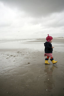Niederlande, Rotterdam, kleines Mädchen mit roter Mütze und gelben Gummistiefeln läuft am nassen Sandstrand - SAF000013