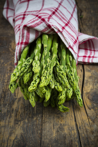 Bündel grüner Spargel, Asparagus officinalis, eingewickelt in ein Küchentuch, auf dunklem Holz liegend, lizenzfreies Stockfoto