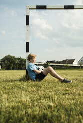 Germany, Mannheim, Teenage boy sitting on grass, leaning on goal - UUF001128