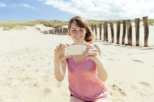 Porträt einer Frau, die mit ihrem Smartphone am Strand fotografiert - MFF001134