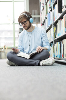 Student in einer Universitätsbibliothek, der mit Kopfhörern auf dem Boden sitzt - WESTF019577