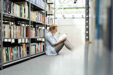 Student in a university library sitting on floor reading book - WESTF019615