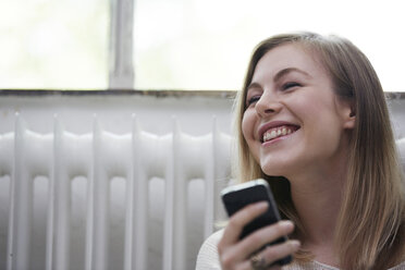 Lachende junge Frau mit Mobiltelefon am Heizkörper - STKF000996