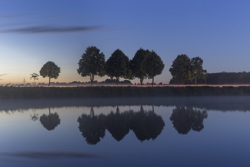 Germany, Lower Saxony, Gifhorn, Lake and reflections of trees, Road with light trail in the evening - PVCF000025