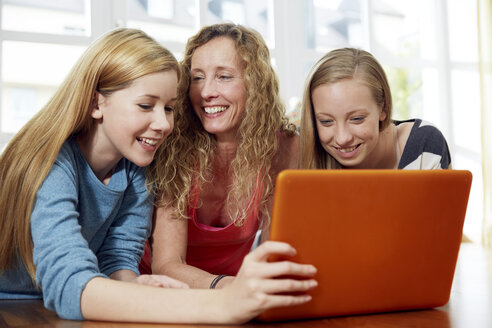 Mother and her two daughters lying on wooden floor at home using laptop - STKF001031