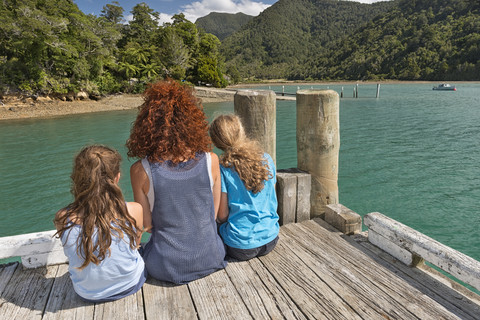 Neuseeland, Südinsel, Marlborough Sounds, Tennyson Inlet, Mutter mit zwei Kindern sitzt auf einem Holzsteg, lizenzfreies Stockfoto