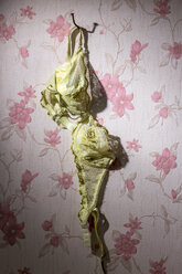 Gelber BH, der an einem gebogenen Nagel hängt, vor einer Tapete mit rosa Blumenmuster - EJWF000408