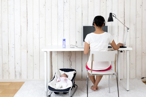 Frau arbeitet im Büro zu Hause, während ihr Baby in einem Schaukelsitz liegt, lizenzfreies Stockfoto