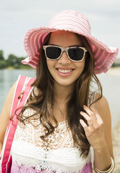 Porträt einer lächelnden jungen Frau mit Sonnenbrille und rosa Hut am Strand - UUF001207