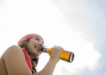 Porträt einer lächelnden jungen Frau mit Bierflasche vor einem bewölkten Himmel - UUF001284