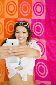 Porträt einer lächelnden jungen Frau, die auf einem Strandtuch liegt und ein Selfie macht - UUF001293