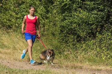 Junge Frau joggt mit Hund auf einem Feldweg - STSF000436