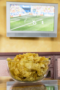 Glasschale mit Kartoffelchips vor einem Flachbildfernseher, auf dem ein Fußballspiel übertragen wird - EJWF000413