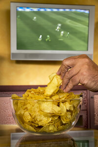 Männerhand nimmt Kartoffelchips aus einer Glasschale vor einem Flachbildfernseher, der ein Fußballspiel überträgt - EJWF000448