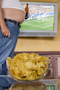 Mann mit nacktem Bierbauch, der eine Bierflasche vor einem Flachbildfernseher hält, der ein Fußballspiel überträgt, Teilansicht - EJWF000449