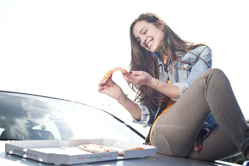 Glückliche junge Frau isst Pizza auf der Motorhaube - FEXF000122
