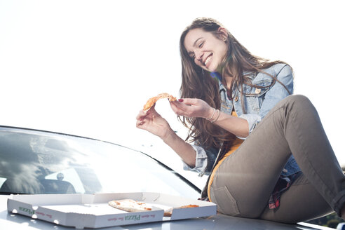 Happy young woman eating pizza on car hood - FEXF000122