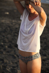 Woman wearing bikini pants and wet t-shirt standing on the beach at twilight - FAF000044