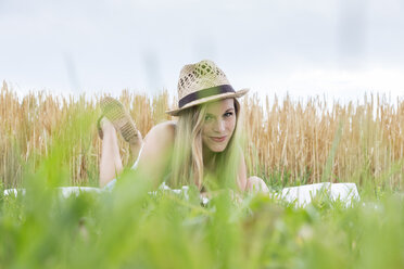 Portrait of smiling young woman with hat lying on a blanket in front of a rye field - DRF000967