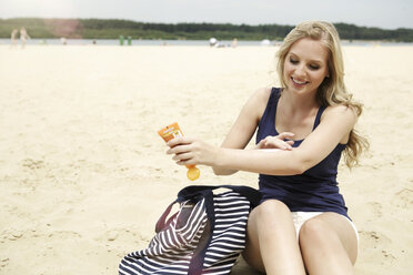 Lächelnde junge Frau, die am Strand sitzt und sich mit Sonnencreme eincremt - GDF000356