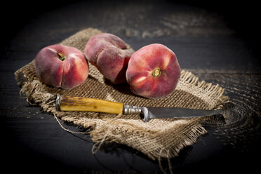 Three doughnut peaches, Prunus persica var. platycarpa, and a knife on jute and dark wooden table - MAEF008808