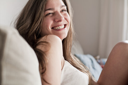 Porträt einer lächelnden jungen Frau, die auf der Couch in ihrem Wohnzimmer sitzt - FEXF000152