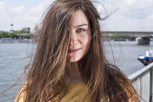 Germany, Cologne, portrait of smiling young woman with blowing hair standing in front of Rhine River - FEXF000215