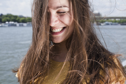 Deutschland, Köln, Porträt einer lächelnden jungen Frau mit wehendem Haar vor dem Rhein, lizenzfreies Stockfoto