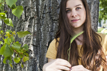 Portrait of young woman with spike in her hand sitting in front of tree trunk - FEXF000220