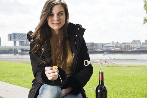 Deutschland, Köln, Porträt einer lächelnden jungen Frau mit Rotweinflasche vor dem Rhein sitzend - FEXF000230