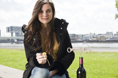 Germany, Cologne, portrait of smiling young woman with red wine bottle sitting in front of Rhine River - FEXF000230