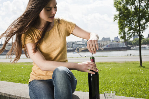 Deutschland, Köln, junge Frau sitzt vor dem Rhein und öffnet eine Rotweinflasche - FEXF000221