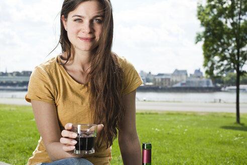 Deutschland, Köln, Porträt einer lächelnden jungen Frau mit einem Glas Rotwein vor dem Rhein sitzend - FEXF000222