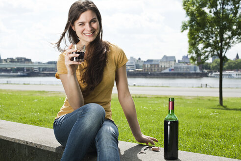 Germany, Cologne, portrait of smiling young woman sitting in front of Rhine River drinking red wine - FEXF000224